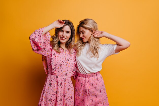 Glamorous woman in cute pink dress spending leisure time with sister. Indoor photo of amazing female models expressing positive emotions.