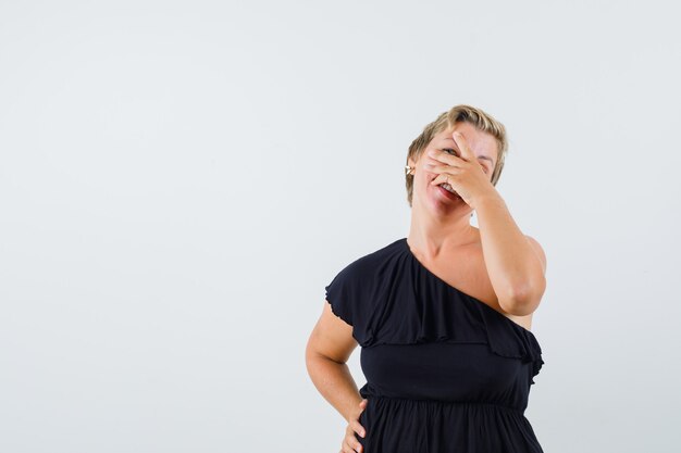 Glamorous woman holding hand on face while looking through fingers in black blouse 