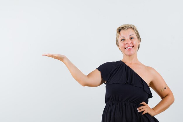 Glamorous woman spreading her hand with open palm aside in black blouse and looking glad. front view.