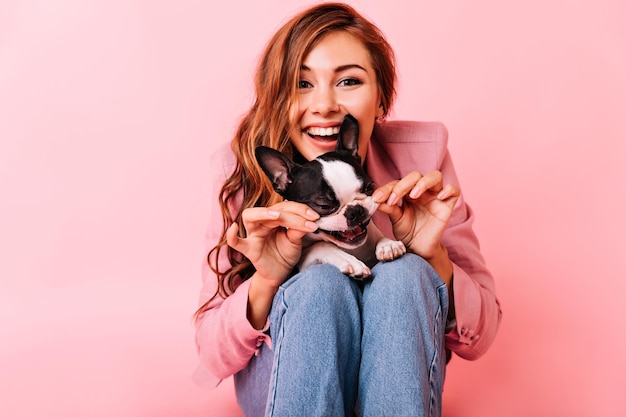 Glamorous woman touching her dogs cheek Indoor photo of blissful female model sitting on pink background with puppy