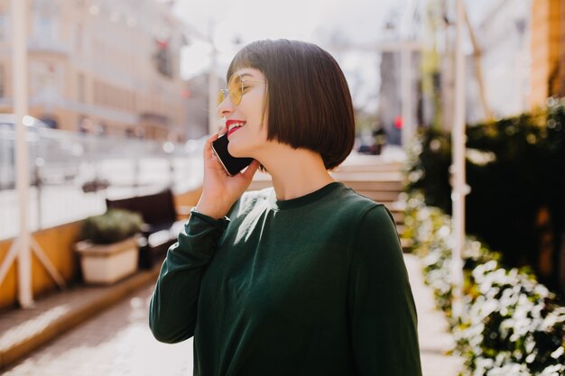 Glamorous woman with short haircut talking on phone. Beautiful brunette girl in green sweater calling someone on street.