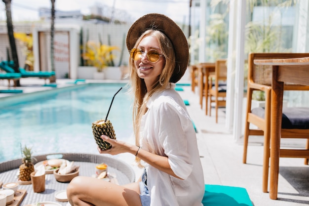 파인애플 칵테일을 마시는 동안 어깨 너머로보고 매력적인 젊은 여자. 과일과 함께 수영장 근처에 앉아 모자에 웃는 금발 소녀.