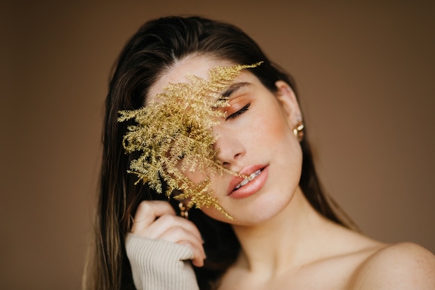 Гламурная молодая женщина позирует с закрытыми глазами на коричневой стене. Добродушная милая дама держит растение.