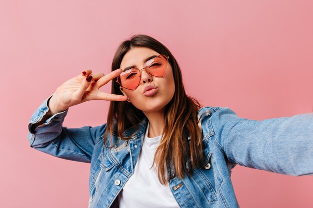 Glamorous young woman taking selfie on pink background. Appealing brunette girl showing peace sign.