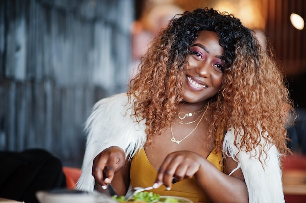 Гламурная африканская американка в желтом платье и белой шерстяной накидке позирует сидя в ресторане и ест салат