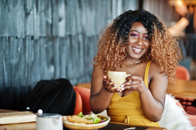Glamour african american woman in yellow dress sitting on restaurant with cup of tea