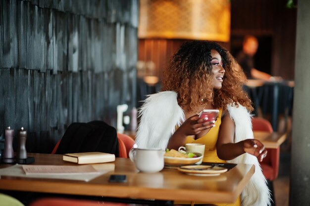 노란색 드레스를 입은 매력적인 아프리카계 미국인 여성이 손에 휴대전화를 들고 레스토랑에서 요리를 들고 테이블에 앉아 있습니다.