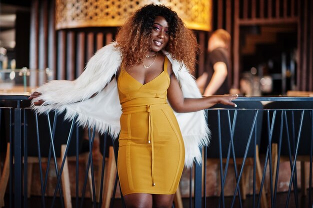 Glamour african american woman in yellow dress and white woolen cape posed at restaurant