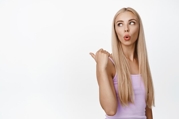 Glamour blond girl in tank top pointing finger left and looking behind her shoulder with surprised face expression white background