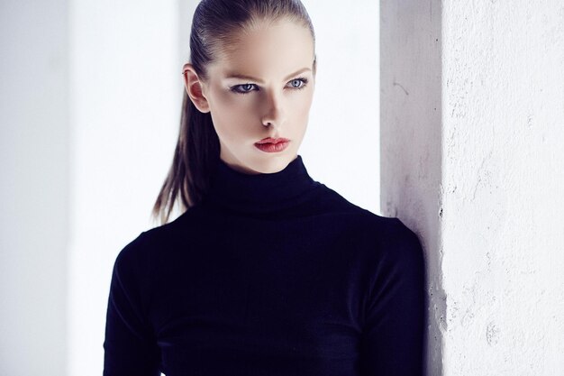 Glamour female model with short brown hair in black dress posing near light wall.