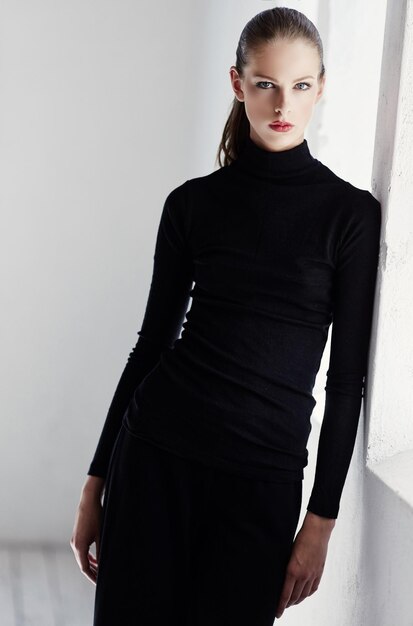 밝은 벽 근처에서 포즈를 취하는 검은 드레스에 짧은 갈색 머리를 가진 매력적인 여성 모델.