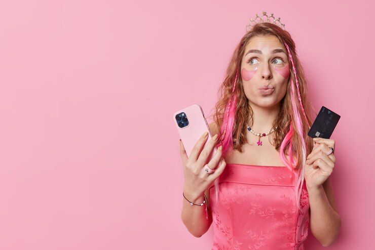 Free Photo glamour feminine woman keeps lips folded pays with credit card for purchase thinks what to buy in online store scrolls eshop applies beauty patches wears crown and dress isolated over pink wall