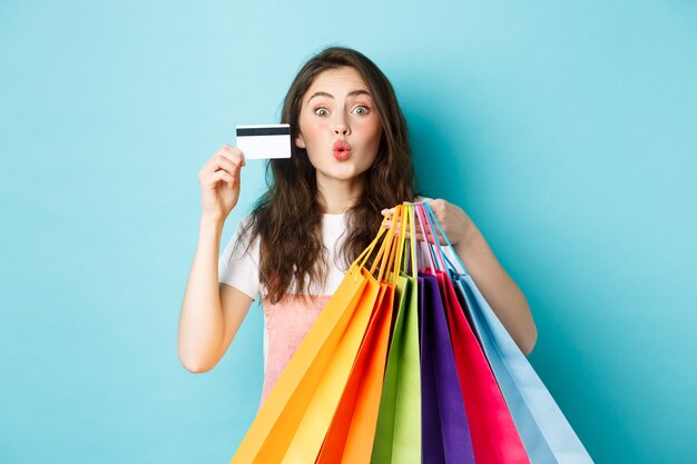 Очаровательная девушка показывает пластиковую кредитную карту и хозяйственные сумки, сморщивает губы для поцелуя, стоя счастливым на синем фоне. Копировать пространство