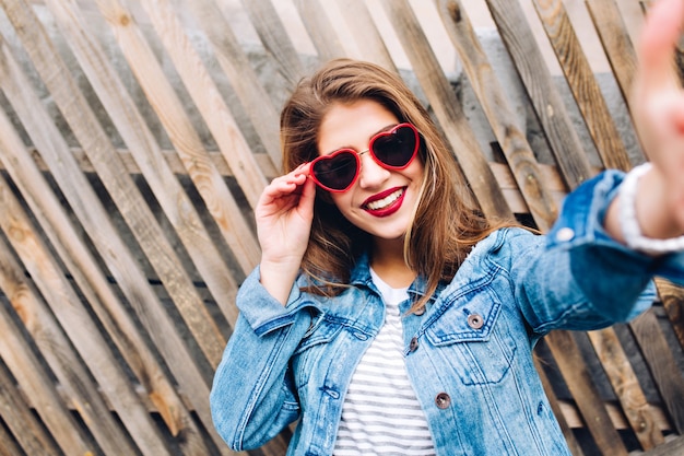 Glamour smiling girl wearing heart glasses holding frame. Close-up portrait of charming young pretty woman touching camera.