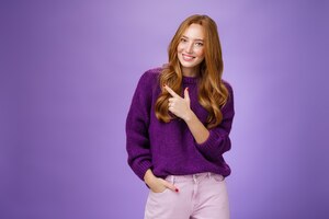 Glamour and stylish feminine woman with long natural red hair in purple sweater holding hand in pocket as pointing at upper left corner showing place she does hairstyle or makeup over violet wall.