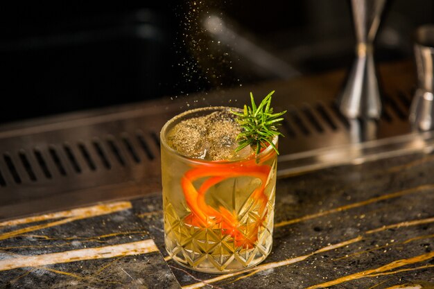 A glass of alcohol cocktail with rosemary leaves, ice cubes and orange spaghetti skin 