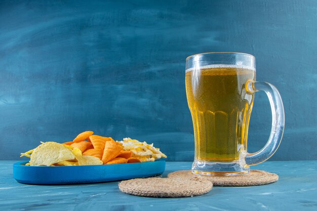 Free photo glass of beer on a trivet next to various chips in a wooden plate , on the blue background.