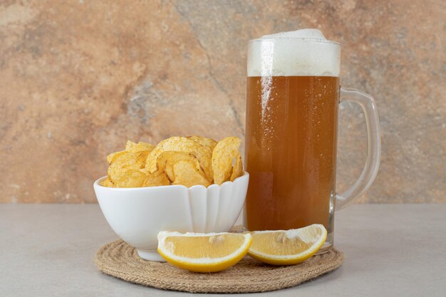 Стакан пива с дольками лимона и миска чипсов на каменном столе