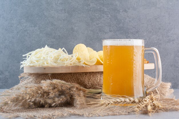 A glass of beer with wheat and potato chips on hay.