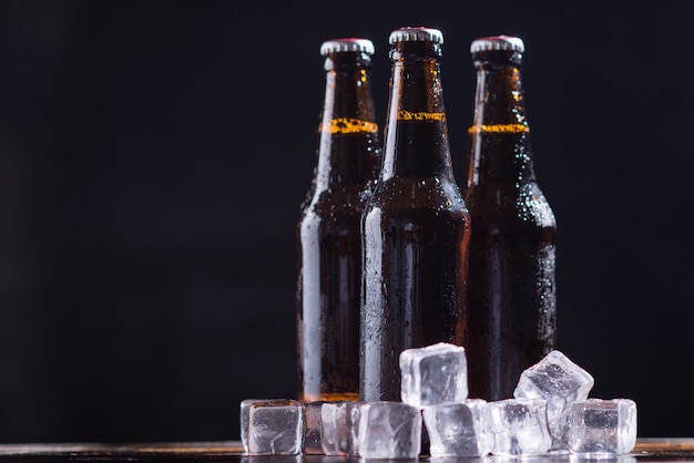 Glass bottles of beer with glass and ice on dark background