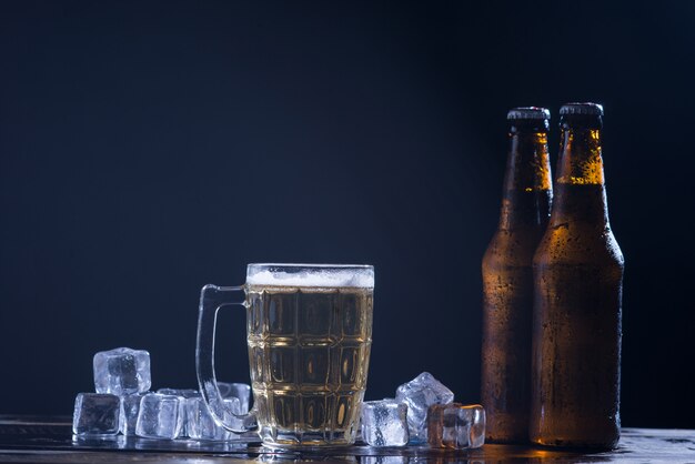 Glass bottles of beer with glass and ice on dark background