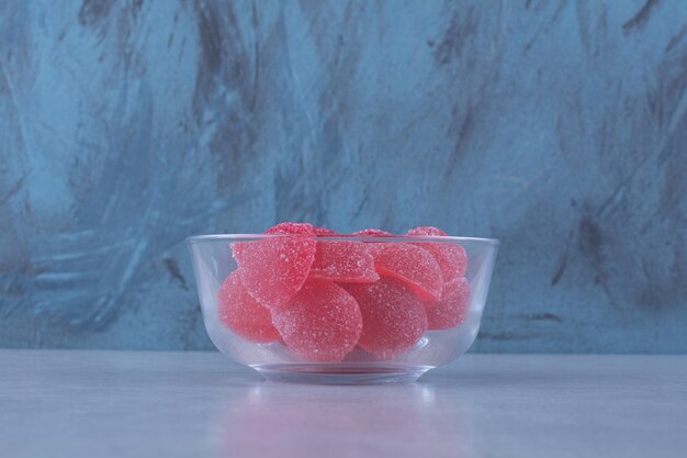 A glass bowl full of red sugary jelly candies on gray table . 