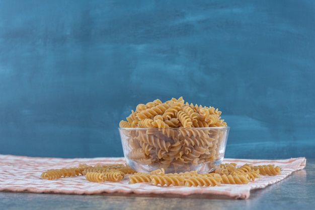 A glass bowl of fusilli pasta, on the marble background.