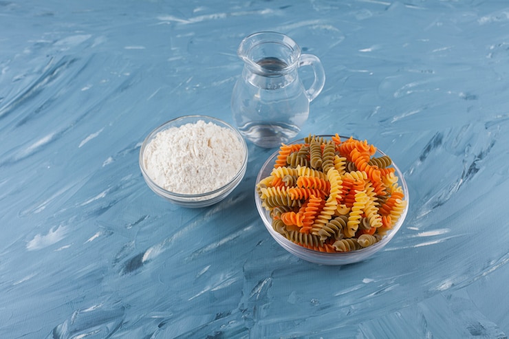 मुफ़्त फोटो a glass bowl of multi colored raw spiral pasta with a pitcher and flour .