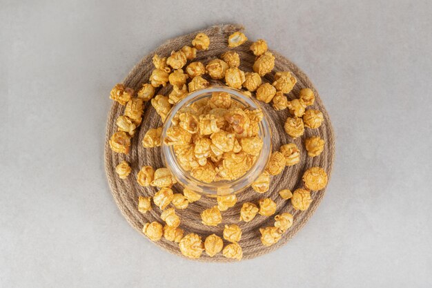 Glass bowl placed on a knitted trivet, filled and encircled with caramel coated popcorn on marble table.