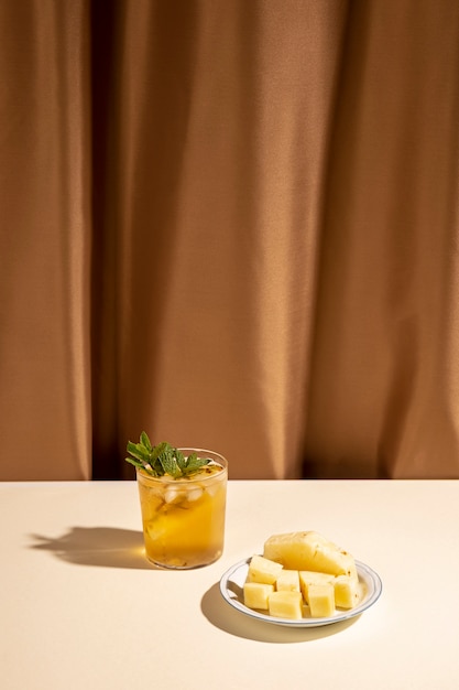 Glass of cocktail drink with pineapple slices on plate over white table against brown curtain