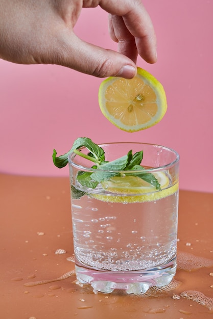 A glass of cold refreshing lemonade on pink surface and woman holding a slice of lemon