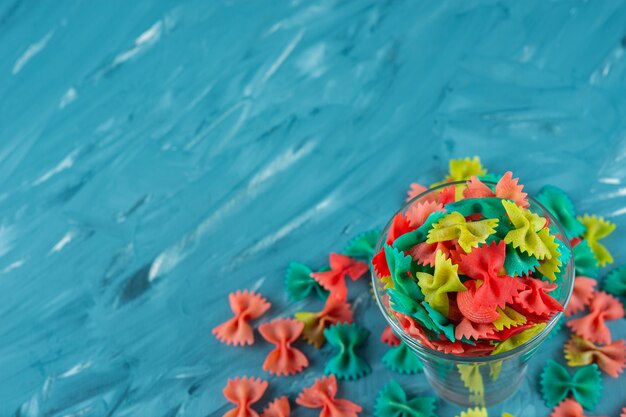 Glass of colorful raw farfalle pasta on blue background. 