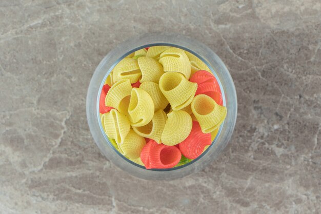 Glass of colorful seashell pasta on marble surface.