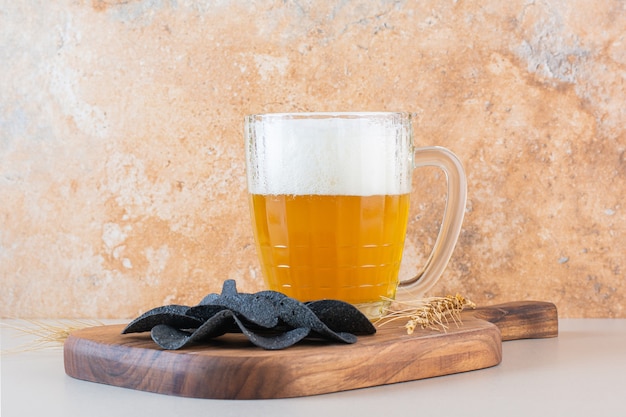 A glass cup of beer with dark potato chips on white background .