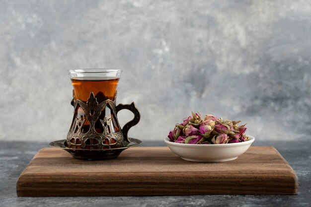 A glass cup of hot tea with dried roses on a wooden cutting board . 