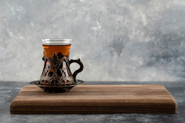 A glass cup of hot tea on a wooden cutting board . 