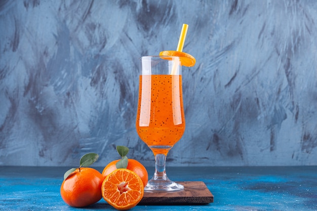 A glass cup of juice with straw and whole and sliced tangerines.