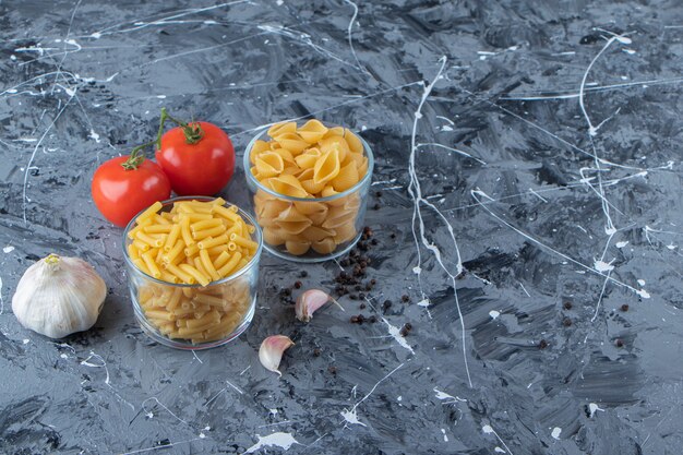 大理石の背景に2つのフレッシュトマトとニンニクが入った生パスタのガラスカップ。