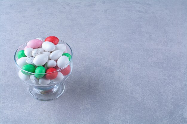 A glass deep plate full of colorful bean candies on gray table. 