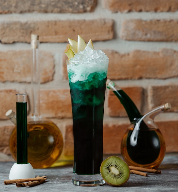 A glass of green alcohol cocktail with apple slices and ice cubes.