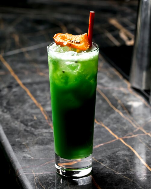 A glass of green cocktail garnished with dried orange and red paper straw