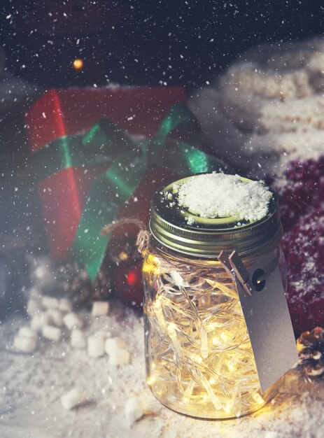 Стеклянная банка с огнями с подарком рядом с ним в то время как снег идет смотреть сверху