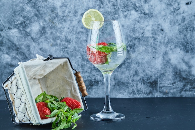 A glass of juice with whole fruits inside and basket of strawberries on blue surface