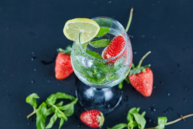 A glass of juice with whole fruits and slice of lemon inside on dark surface