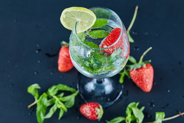 A glass of juice with whole fruits and slice of lemon inside on dark surface