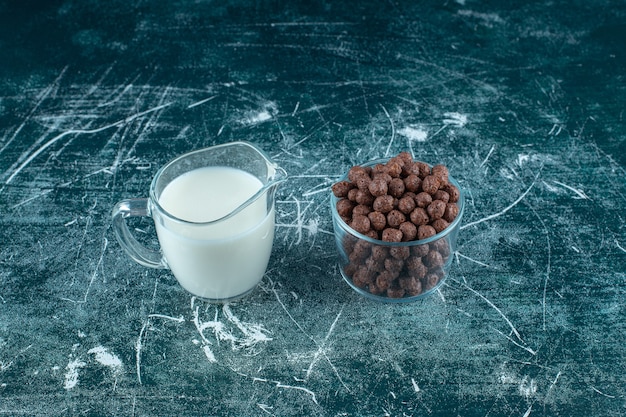 A glass of milk next to corn balls in a glass bowl on the blue surface