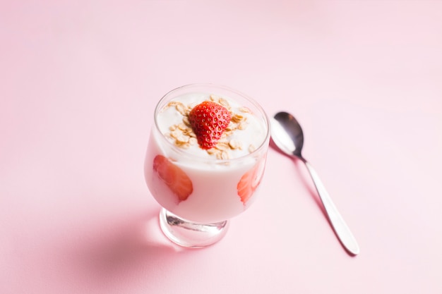 The glass of oat granola with yogurt and fresh strawberries and spoon on pink background