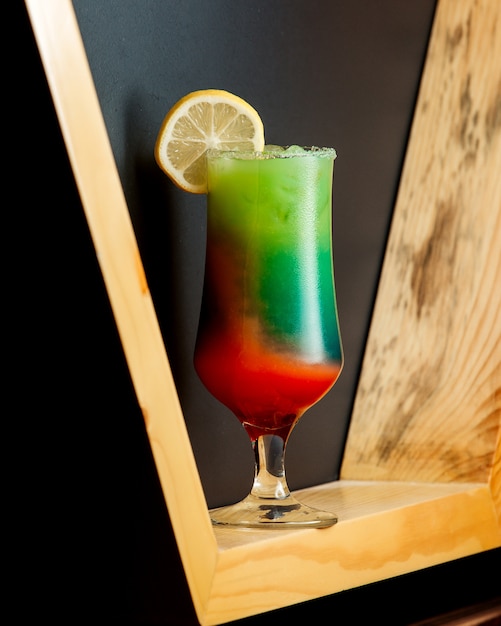 A glass of ombre cocktail with green and orange colors topped with lemon