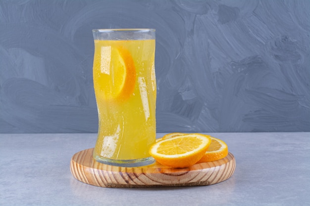 Free photo a glass of orange juice next to slice orange on a wooden plate on marble table.