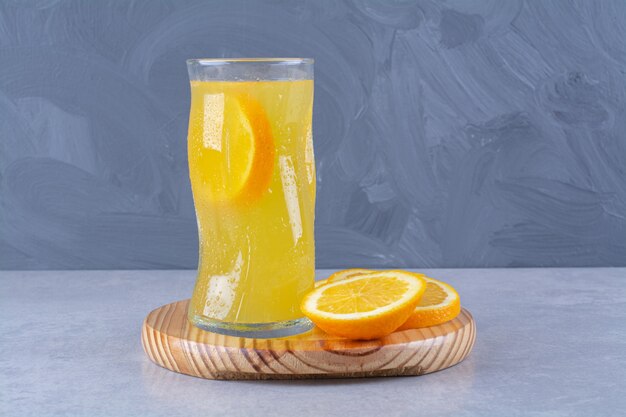 A glass of orange juice next to slice orange on a wooden plate on marble table.